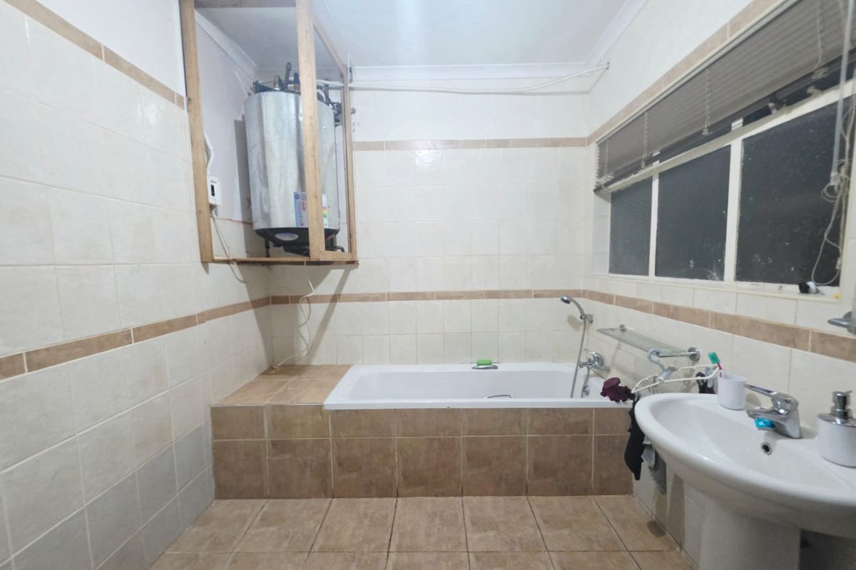 A functional bathroom featuring a built-in bathtub, pedestal basin, and ceramic tiling, currently requiring cosmetic updates and enclosure of the exposed water heater unit.