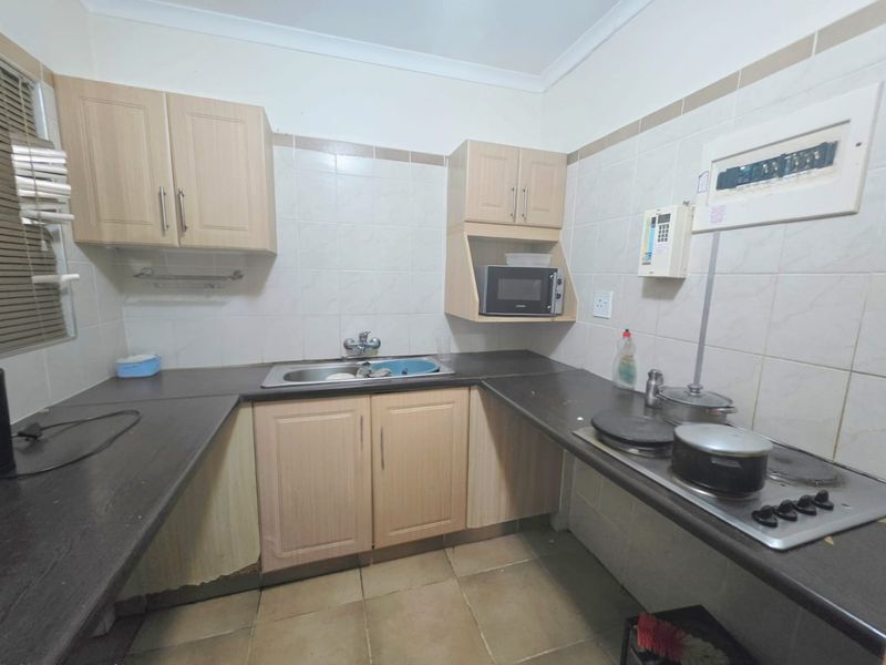 A compact kitchen featuring light wood-finish cabinetry, dark countertops, a built-in electric hob, and tiled splashbacks, requiring some maintenance to the lower cabinet base.
