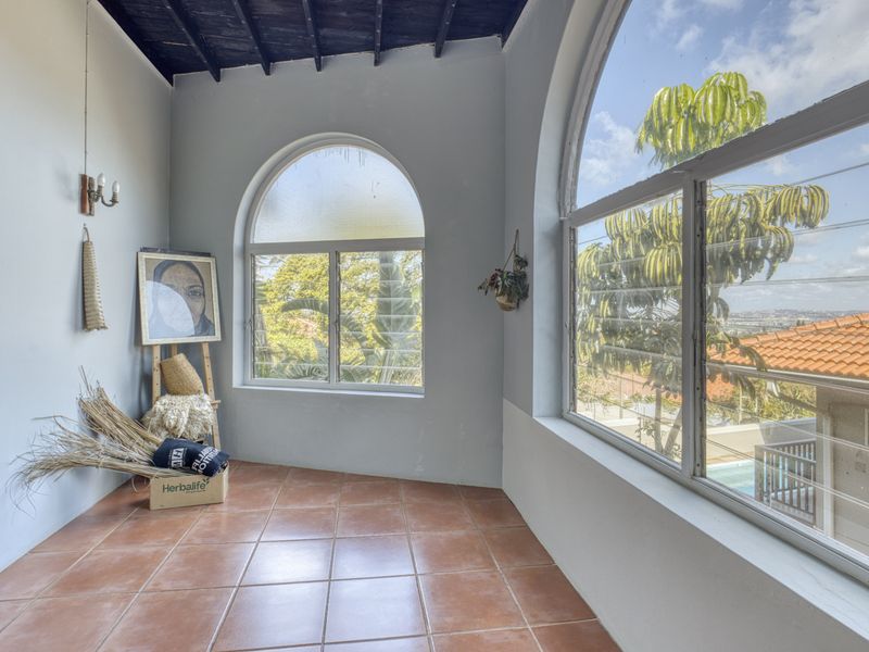 A bright, enclosed patio or sunroom featuring terracotta floor tiles, exposed timber ceiling beams, and large arched windows with views.