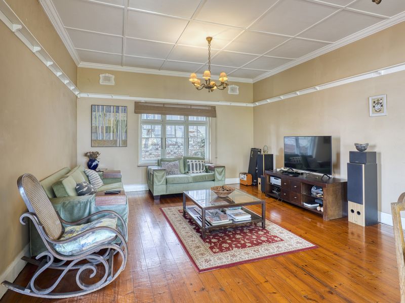 A spacious living room featuring original hardwood flooring, a classic coffered ceiling, and a large multi-pane window providing natural light.