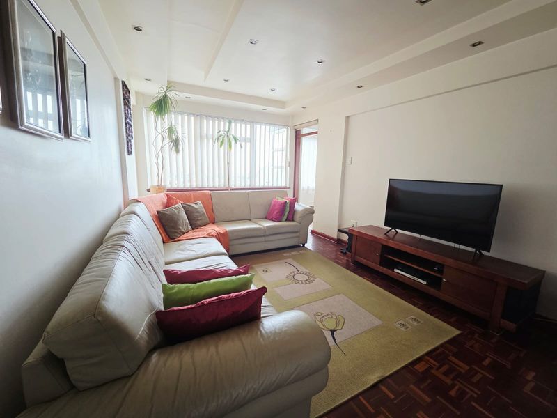 A comfortable living room featuring classic parquet flooring, recessed ceiling lighting, and large windows with vertical blinds, offering a bright and functional space.