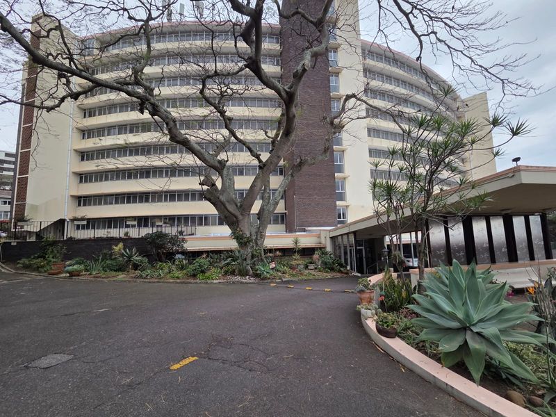 A view of the front exterior of a multi-story apartment building featuring a curved facade, brick accents, and a paved driveway entrance.