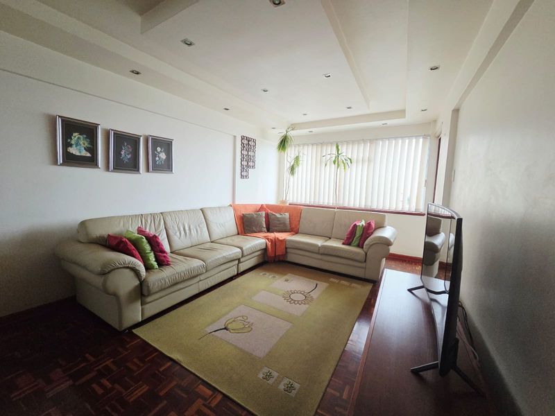 A well-lit living room featuring parquet flooring, recessed ceiling lighting, and large windows with vertical blinds.