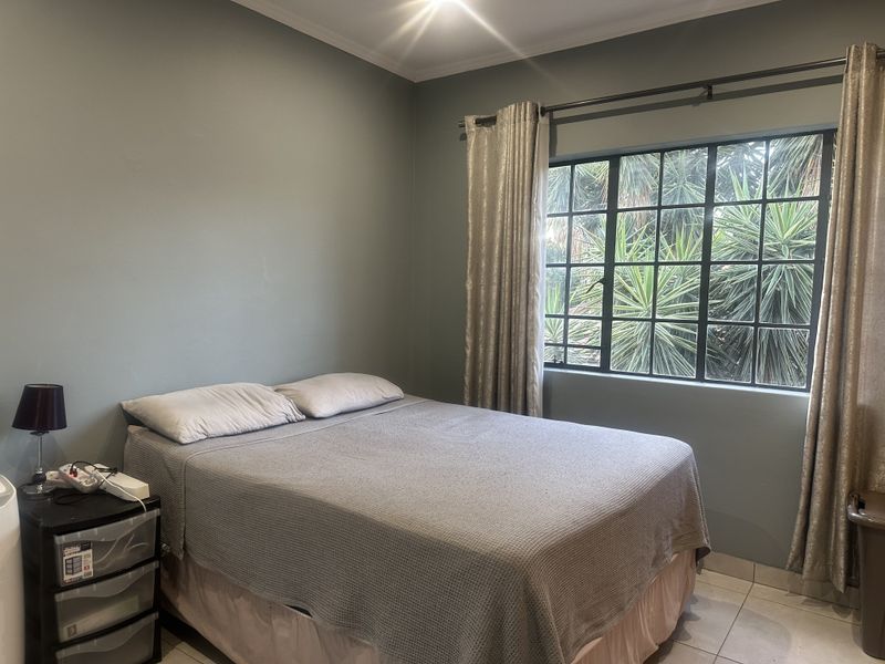 A functional bedroom featuring tiled flooring and a large steel-framed window with curtain fixtures, offering a view of the exterior greenery.