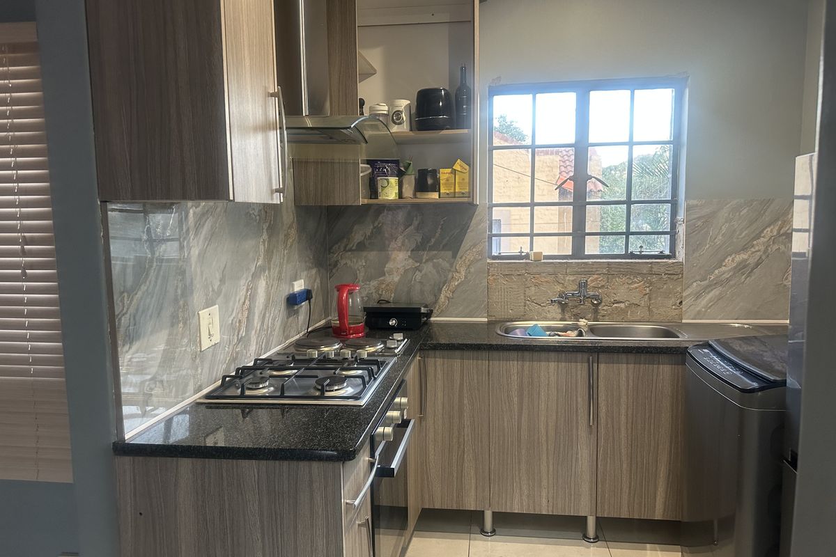 Compact kitchen featuring wood-finish cabinetry, granite countertops, gas hob, extractor fan, and marble-look splashback tiles, with some minor finishing work required around the window.