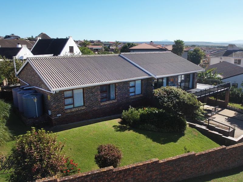 Aerial view of a brick-faced house featuring a tiled roof, raised wooden deck, and water storage tanks, set within a well-maintained garden.