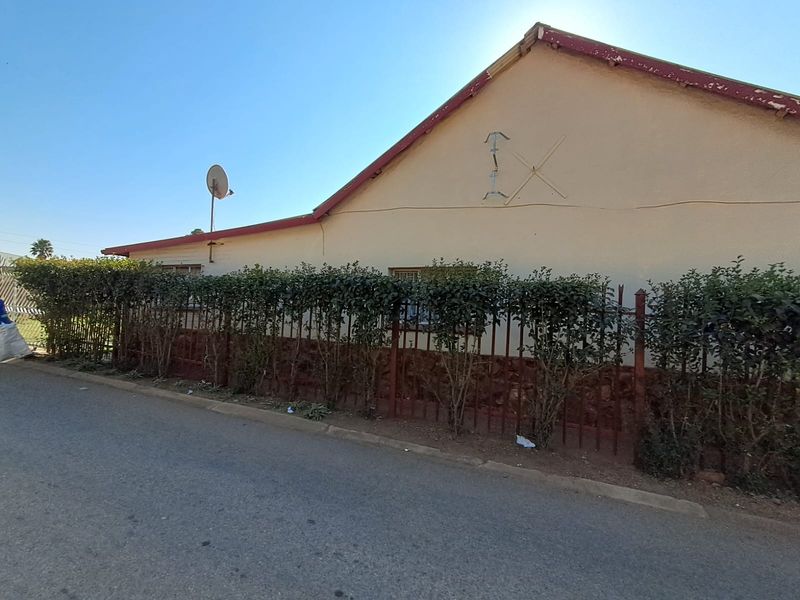 Street-facing view of the property exterior featuring a gabled roof, perimeter metal fencing, and established hedge screening.