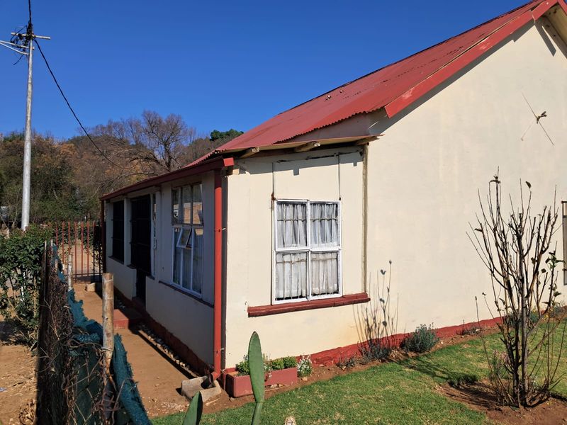 Side view of a residential house featuring a red corrugated metal roof, plastered walls, and traditional casement windows with security bars.