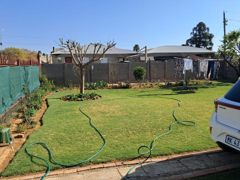 A private backyard featuring a well-maintained lawn, established garden beds, a precast concrete boundary wall, and a shade sail structure.