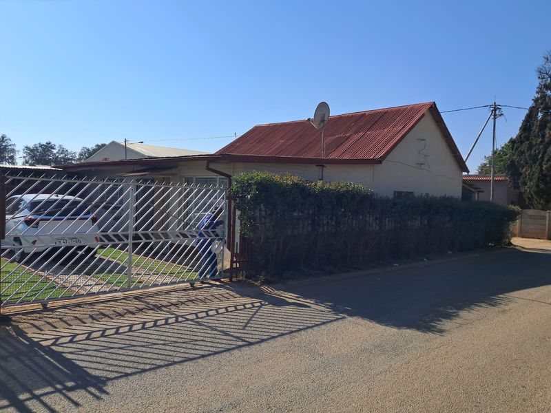 Front exterior view of a single-story house featuring a corrugated metal roof, perimeter hedge, and a metal security gate.