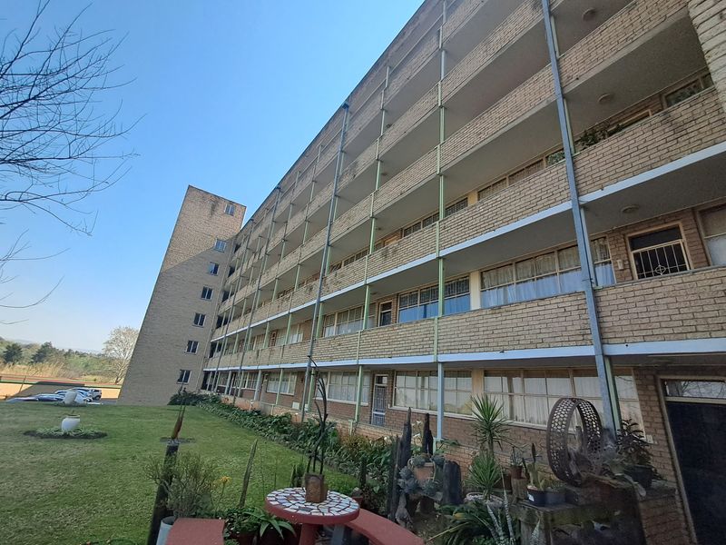 Exterior view of a multi-story apartment building featuring a brick facade, continuous balconies, and metal structural supports overlooking a garden area.