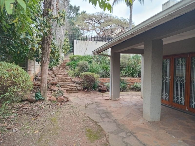 A covered patio area featuring stone paving and concrete pillars, leading to a landscaped garden with stone stairs and a retaining wall.