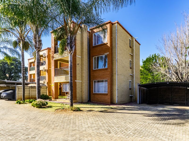 Front view of a multi-story brick apartment building featuring covered parking carports, paved driveway, and established palm trees.