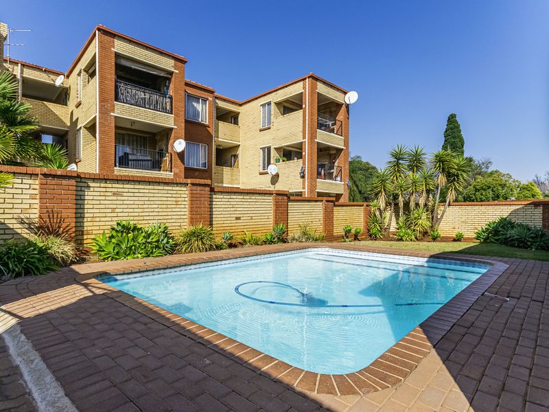 A well-maintained communal swimming pool area featuring paved decking and a brick boundary wall, set against the backdrop of a multi-story apartment building.