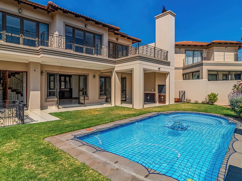 Rear view of a double-story residence featuring a swimming pool, covered patio with built-in braai, and an upper-level balcony with wrought iron railings.