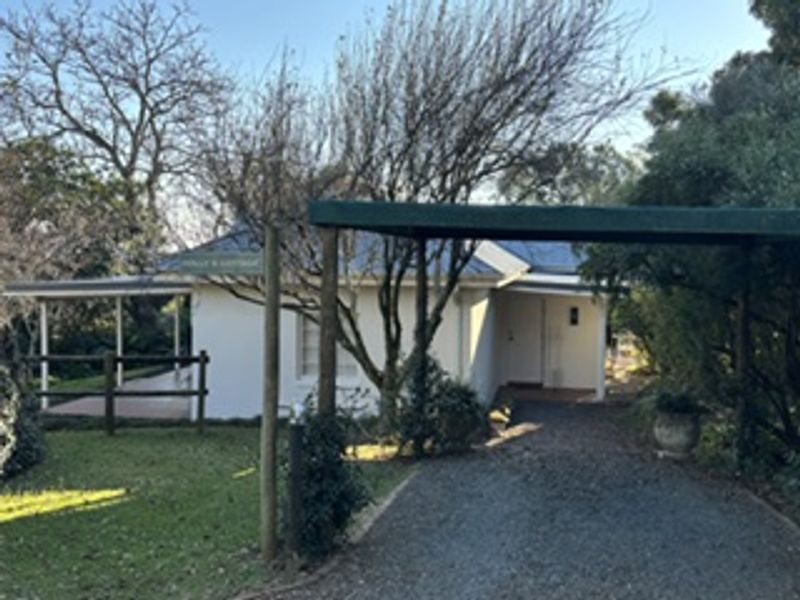 Front view of the cottage featuring a gravel driveway, attached carport, and a covered patio area surrounded by established trees and garden.