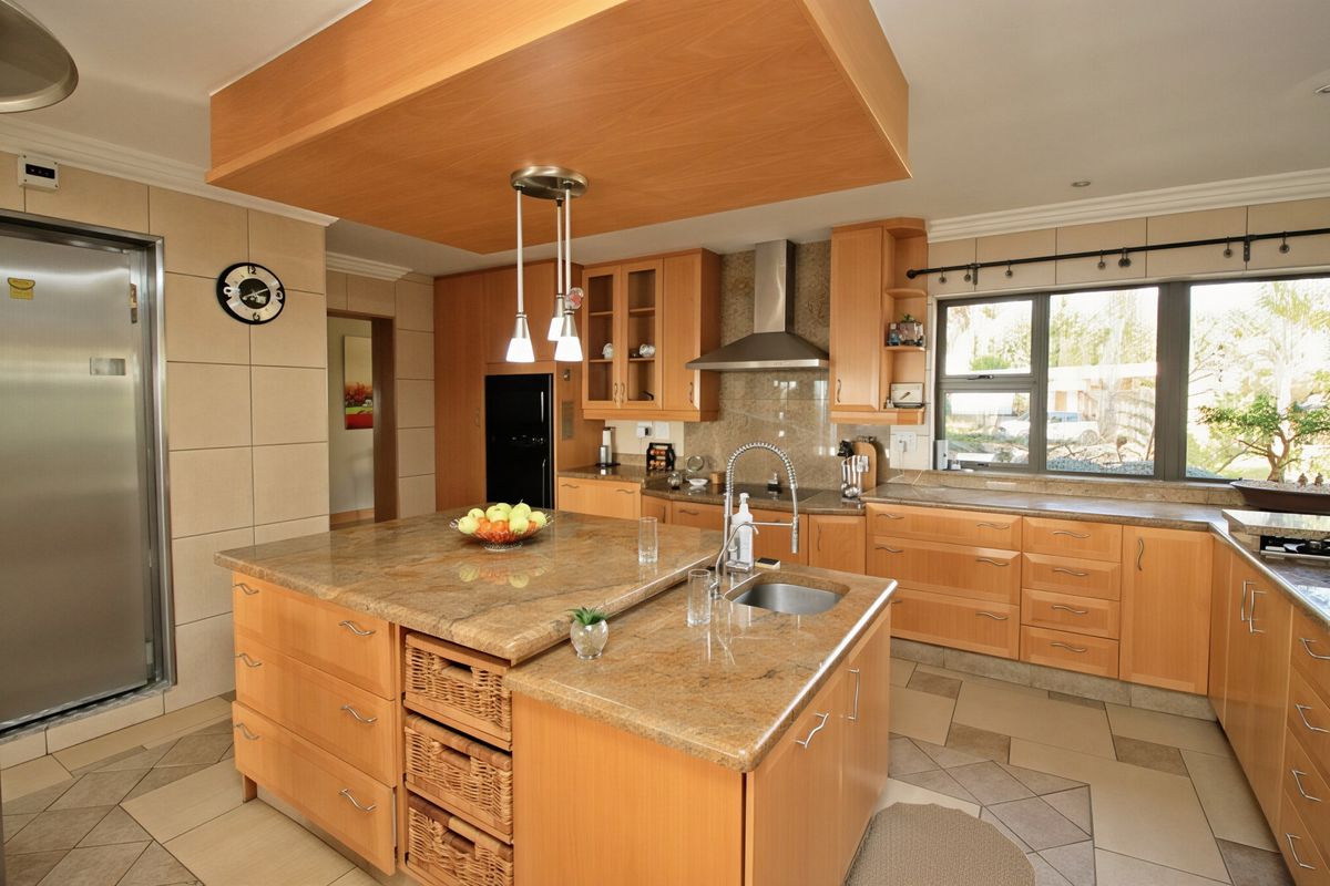 Spacious kitchen featuring warm wooden cabinetry, granite countertops, a central island with integrated sink, and a modern drop ceiling with pendant lighting.