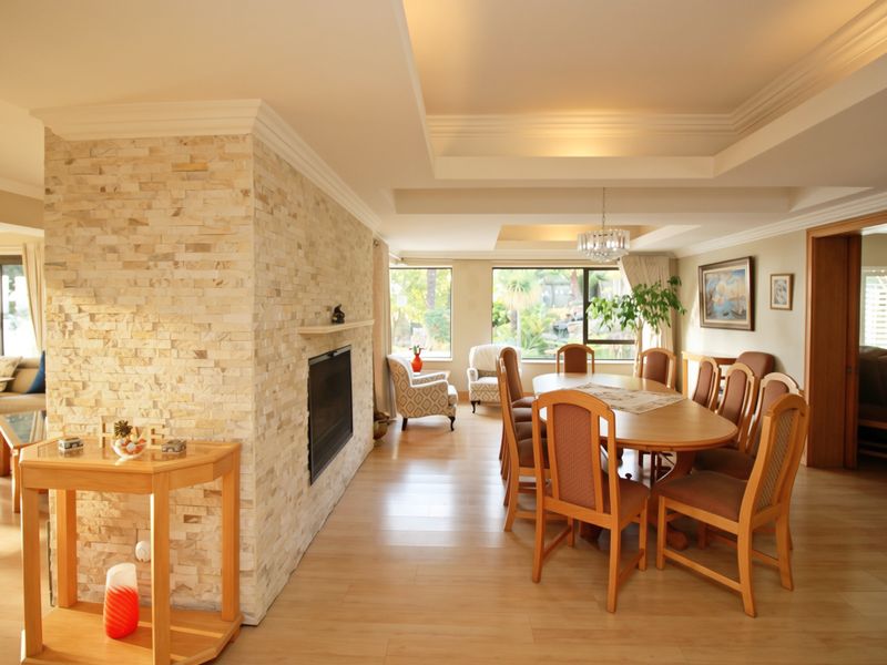 Spacious open-plan living and dining area featuring a prominent stone-clad fireplace, recessed tray ceilings, and light wood-look flooring with natural light.