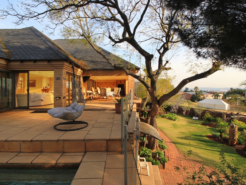 Spacious covered patio featuring tiled flooring and glass balustrades, seamlessly connecting to the garden and pool area with views of the surrounding landscape.