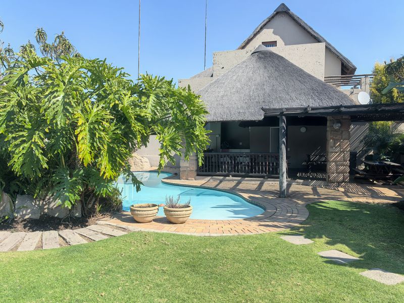 Outdoor entertainment area featuring a swimming pool, a thatched roof lapa with stone pillars, and landscaped garden surroundings.