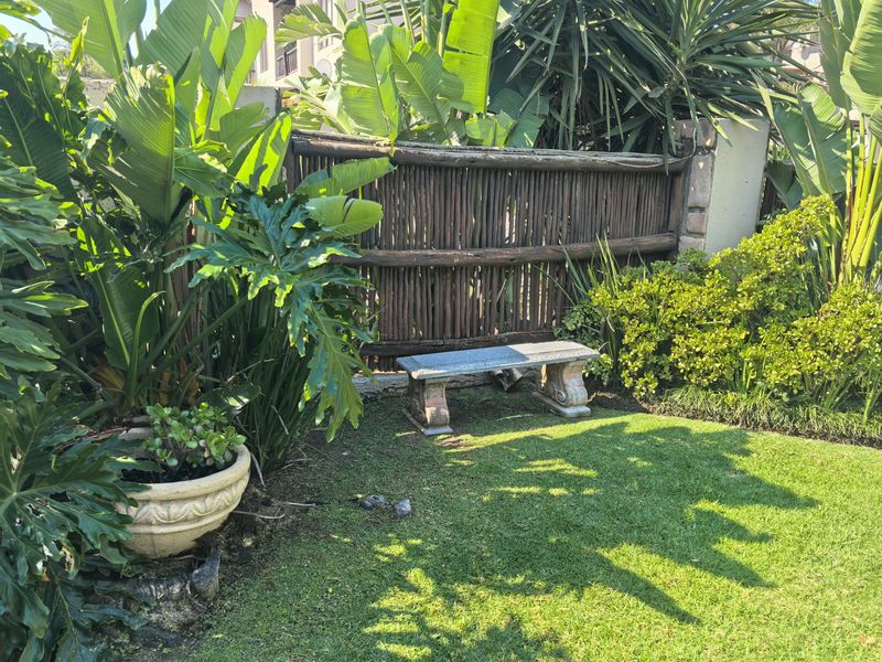 A well-maintained private garden area featuring lush tropical greenery, a natural wooden pole fence, and a stone bench.