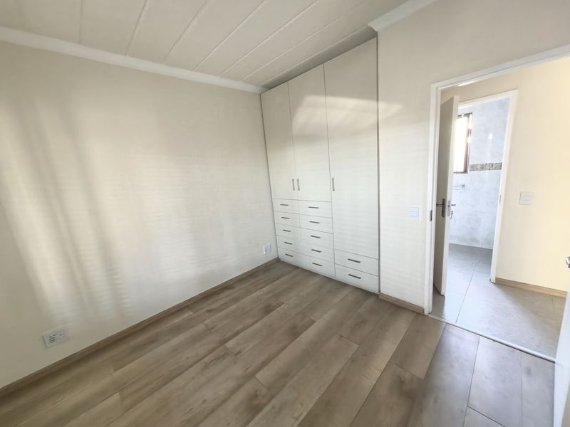 A bright, empty bedroom featuring modern laminate flooring, built-in floor-to-ceiling cupboards, and direct access to an adjoining bathroom.