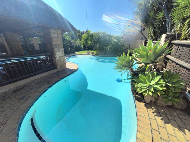 A well-maintained swimming pool area featuring a thatched lapa with stone pillars, surrounded by established garden landscaping and brick paving.