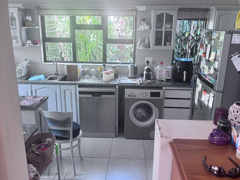 A functional kitchen featuring white cabinetry, granite countertops, tiled flooring, and integrated laundry and dishwasher appliances with a view of the garden.