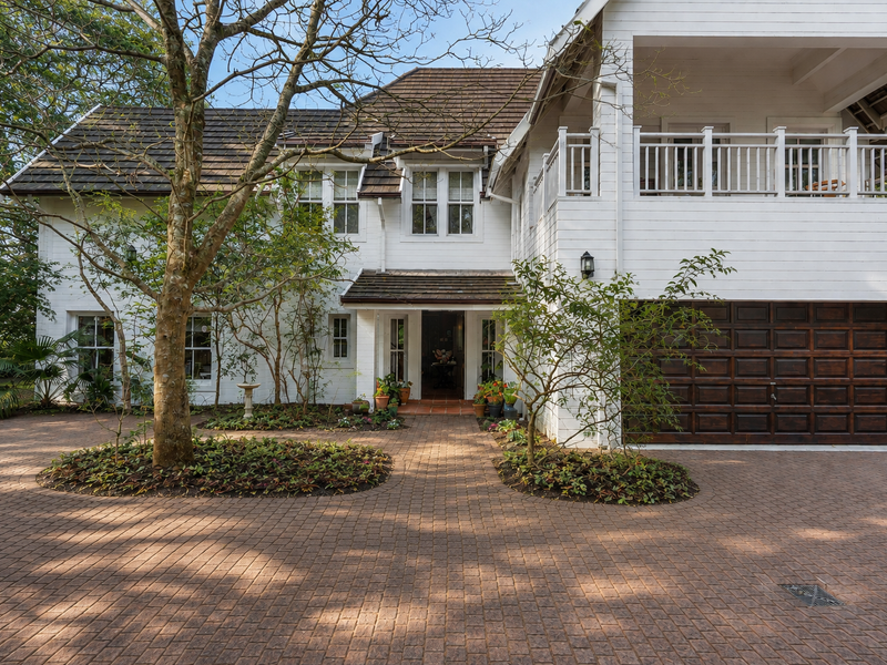 A charming two-story home featuring white timber cladding, a paved driveway, a timber garage door, and an upper-level balcony.