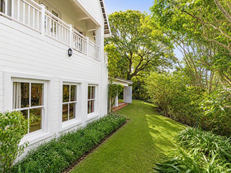 A well-maintained rear exterior view featuring white timber weatherboard cladding, sash windows, and a private upper-level balcony overlooking a lush landscaped garden.