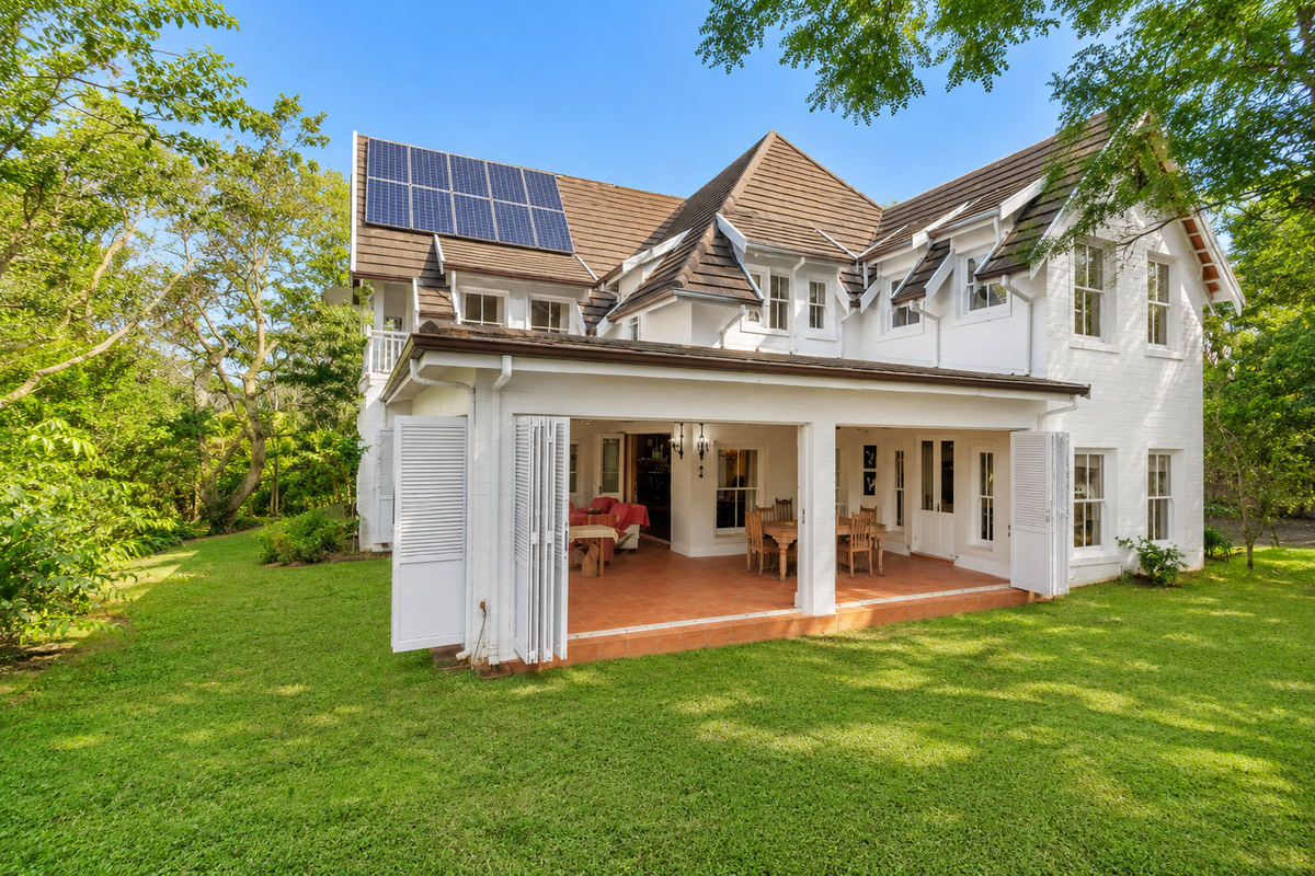 Rear view of a two-story house featuring a covered patio with folding shutters, tiled roof, and installed solar panels set within a lush garden.