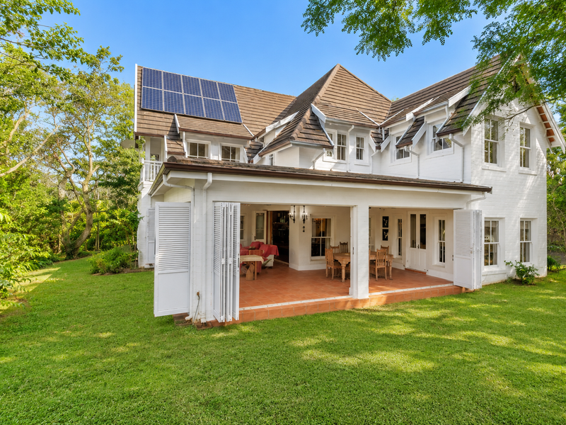 Rear view of a two-story house featuring a covered patio with folding shutters, tiled roof, and installed solar panels set within a lush garden.