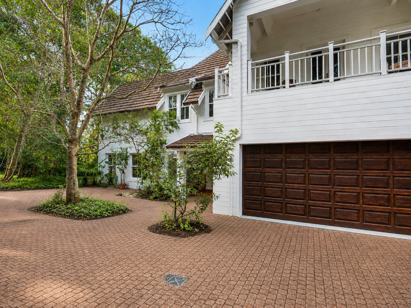 Front elevation of a two-story residence featuring a paved driveway, dark timber garage door, and a white weatherboard facade with an upper-level balcony.