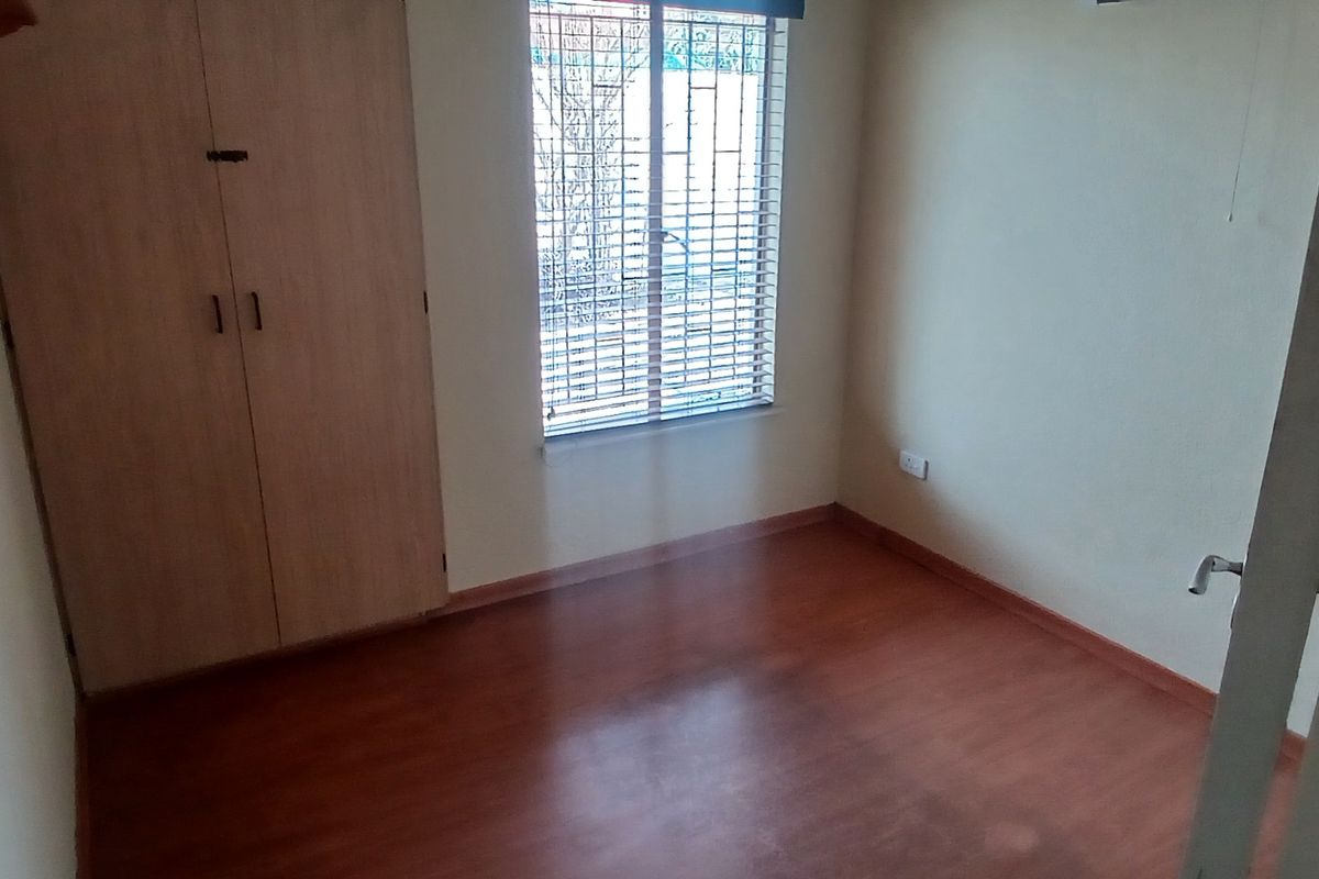 A compact, empty bedroom featuring laminate flooring, a built-in wardrobe, and a window fitted with venetian blinds.