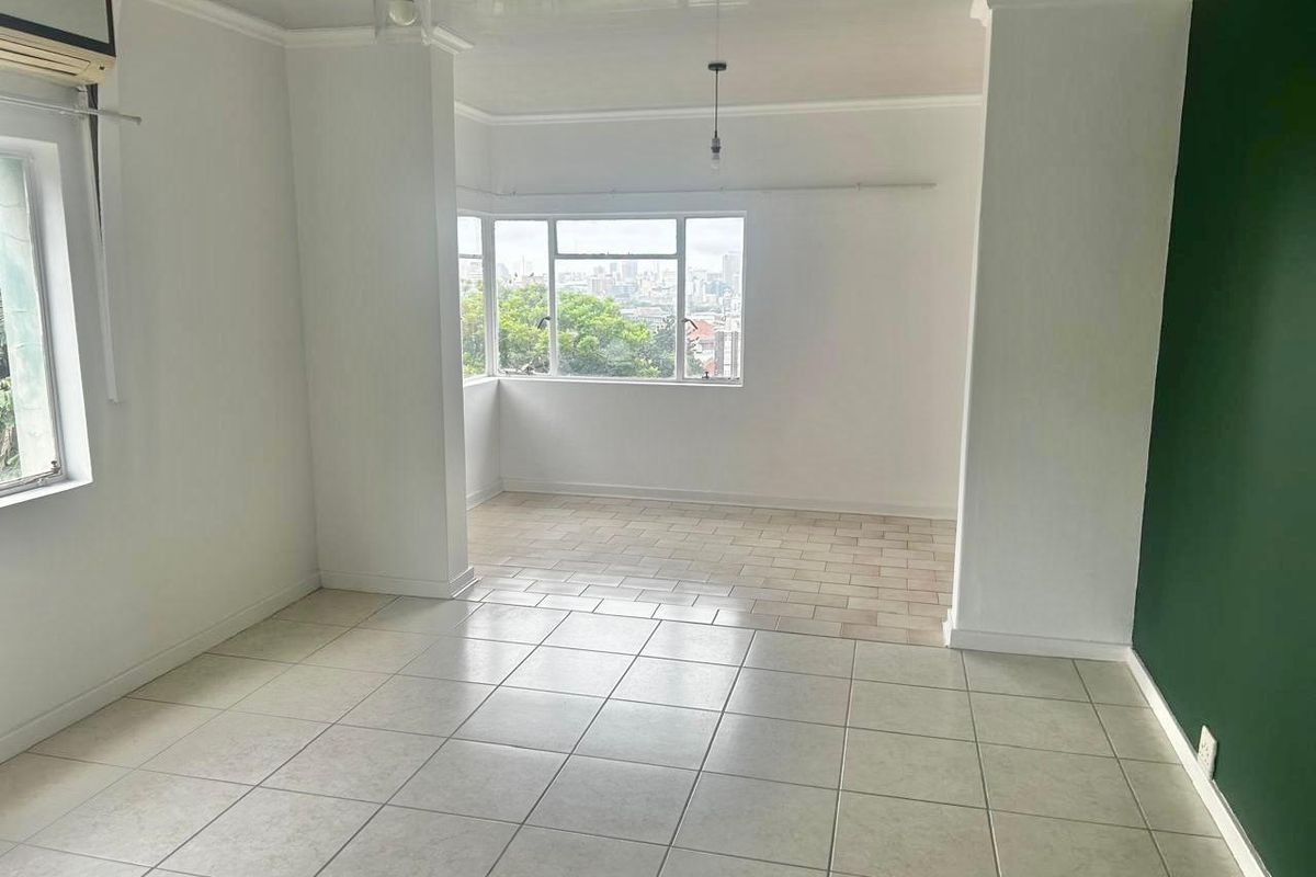 A bright, open-plan living area featuring neutral tiled flooring, large windows with city views, and a modern green accent wall.