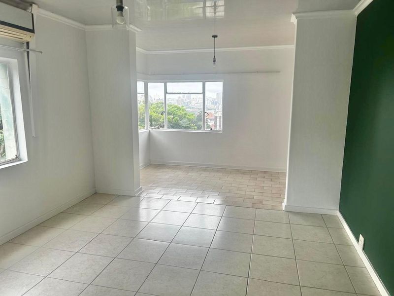 A bright, open-plan living area featuring neutral tiled flooring, large windows with city views, and a modern green accent wall.