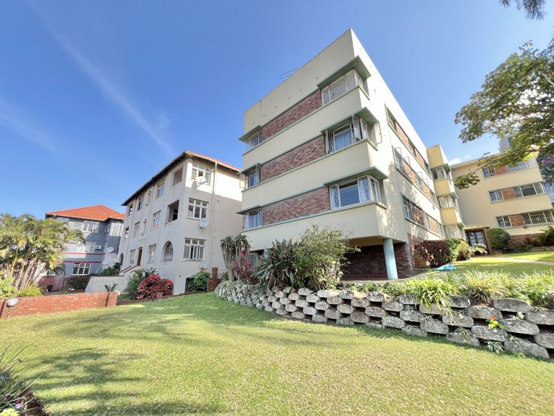 Exterior view of a multi-story apartment building featuring brick and plaster facade, casement windows, and a landscaped garden with a stone retaining wall.