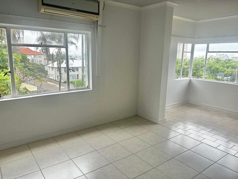 A bright, empty living area featuring tiled flooring, large windows providing natural light, and a wall-mounted air conditioning unit.