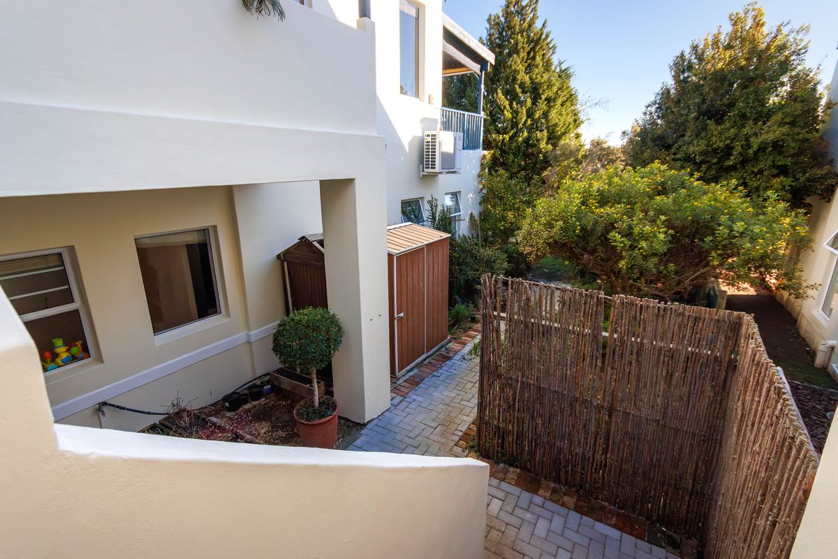 A view of the rear exterior area featuring a paved walkway, established garden greenery, and an external air conditioning unit mounted on the wall.