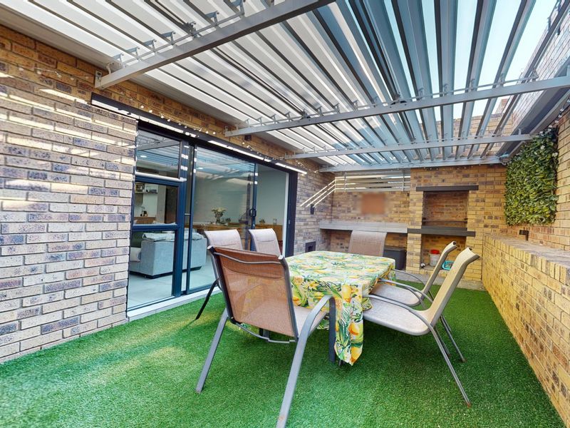 Covered outdoor patio featuring a louvre roof, built-in braai, and artificial grass, providing a functional space for outdoor dining and entertainment.