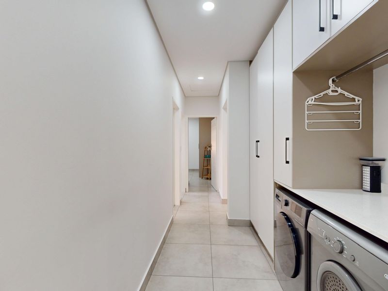 A clean, modern laundry area featuring built-in cabinetry, tiled flooring, and integrated appliance spaces, leading into a bright hallway.