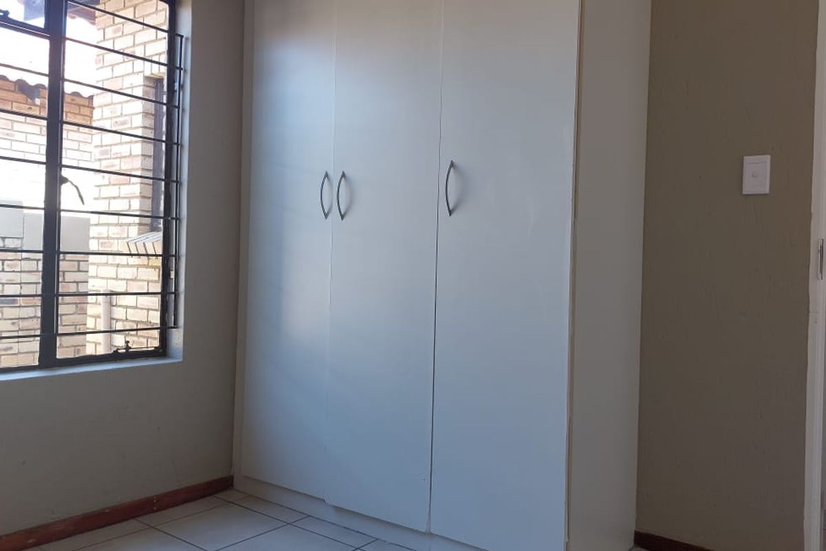 A clean, empty bedroom featuring built-in cupboards, tiled flooring, and a window fitted with security bars.