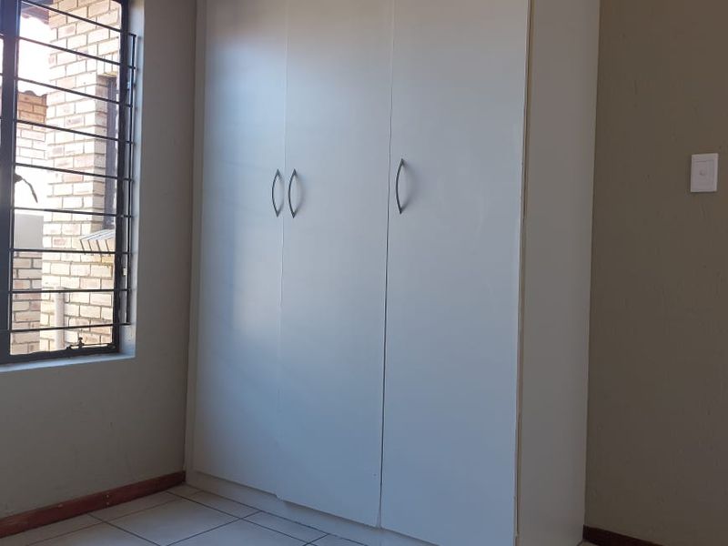 A clean, empty bedroom featuring built-in cupboards, tiled flooring, and a window fitted with security bars.