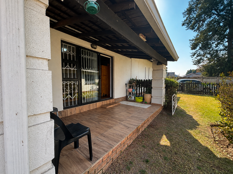 A covered patio area featuring wood-look tiled flooring, exposed timber beams, and a sliding glass door with security gate, overlooking the garden.