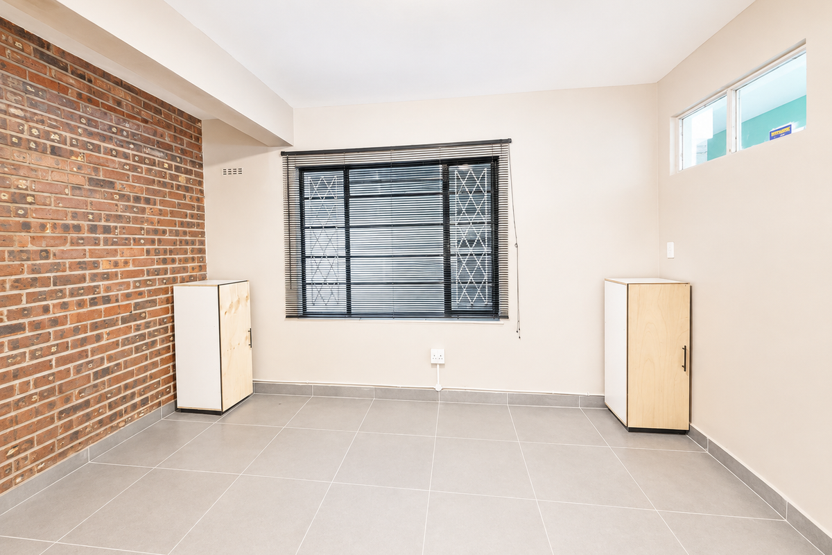 A bright, empty bedroom featuring a striking exposed brick wall, neutral tiled flooring, and secure window treatments with built-in storage units.