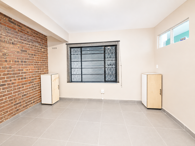A bright, empty bedroom featuring a striking exposed brick wall, neutral tiled flooring, and secure window treatments with built-in storage units.