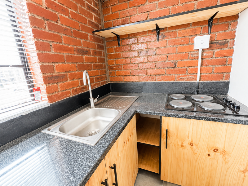 Compact kitchen featuring exposed brick walls, wooden cabinetry, laminate countertops, a stainless steel sink, and a built-in electric hob.