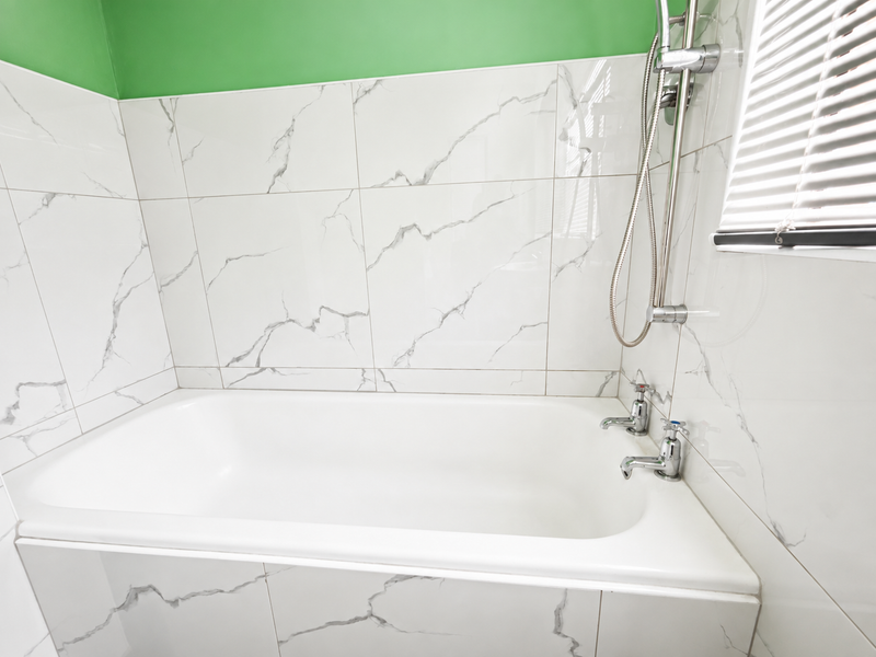 A clean bathroom featuring a built-in bathtub with marble-effect wall tiling, chrome fixtures, and a fitted venetian blind.