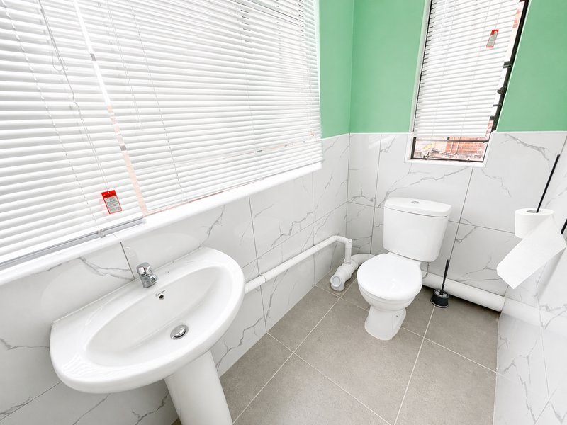 A clean, modern bathroom featuring a pedestal washbasin, toilet, marble-look wall tiling, and neutral floor tiles with functional venetian blinds.
