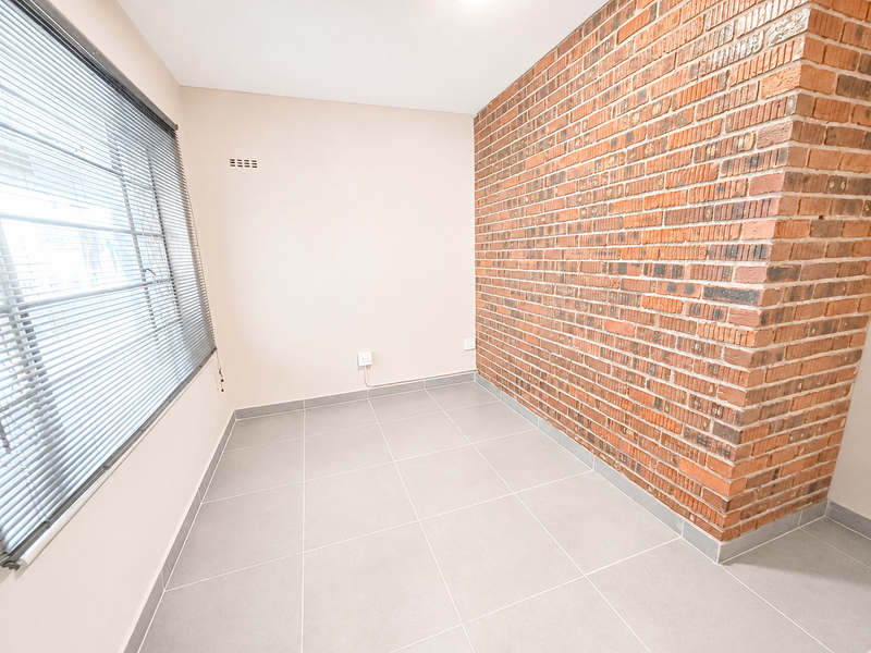 A bright, empty bedroom featuring a prominent exposed brick feature wall, neutral tiled flooring, and fitted venetian blinds.